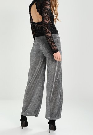 Kvinde iført sort gennemsigtig blonder langærmet top med åben ryg, sølvfarvede bukser med vide ben og sorte ankelstøvler med høje hæle, stående i profil.