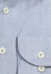 Close-up van een lichtblauw overhemd met visgraatmotief en witte knopen, met nadruk op de stoftextuur en knoopdetails.