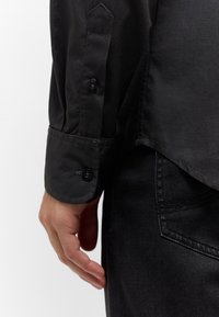Camisa negra con una textura suave, puños abotonados y un dobladillo recto. Llevada con jeans oscuros, mostrando un estilo monocromático.