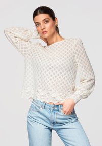 Femme portant un chemisier à manches longues en dentelle blanche et un jean bleu clair, posant avec une main derrière la tête et l'autre dans la poche.