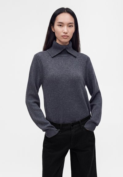 Jeune femme aux longs cheveux noirs portant un pull à col roulé gris côtelé avec col étendu et un pantalon noir, debout les mains dans les poches.