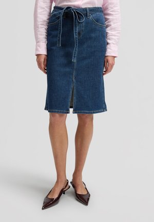 Kvinde iført en knælængde blå denimnederdel med frontslids og bindebælte, kombineret med en lyserød langærmet skjorte og mørke spidse sko.