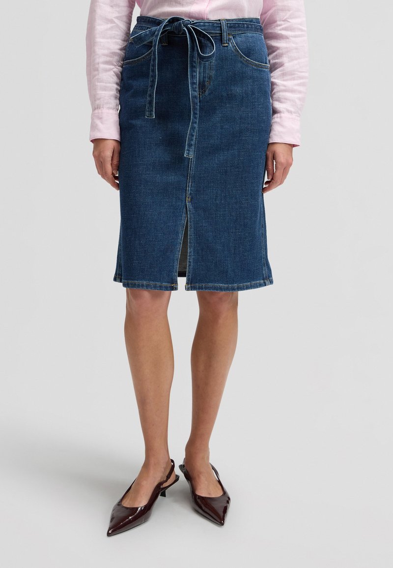 Femme portant une jupe en denim bleue longueur genou avec une fente avant et une ceinture à nouer, associée à une chemise rose clair à manches longues et des chaussures pointues foncées.