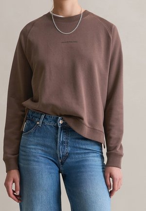 Personne portant un sweat-shirt marron avec le texte "Marc O'Polo Denim" et un jean bleu, accessoirisée d'un collier chaîne en argent.