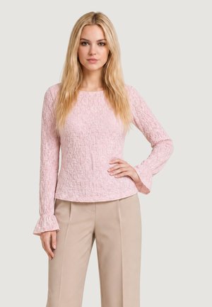 Donna con lunghi capelli biondi che indossa una camicetta rosa chiaro a maniche lunghe con trama e pantaloni beige, in piedi con una mano sull'anca.