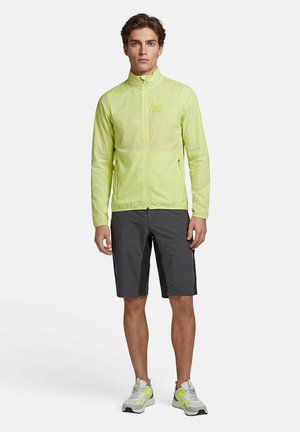 Junger Mann steht, trägt eine hellgrüne leichte Jacke, dunkelgraue knielange Shorts und weiße Sneaker mit limettengrünen Akzenten.