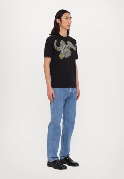 T-shirt en coton noir avec un graphique de tête de taureau en jaune et blanc. Associé à un jean en denim bleu et des chaussures noires.