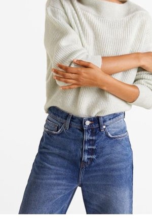 Osoba w kremowym, prążkowanym swetrze i niebieskich, wysokiej talii dżinsach, ze skrzyżowanymi ramionami na wysokości talii.