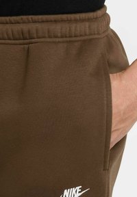 Jogger in cotone marrone con vita elasticizzata, tasca laterale e dettagli del logo Nike bianco. Texture liscia con una silhouette aderente.