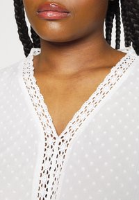 Gros plan d'une personne aux cheveux tressés portant un chemisier blanc avec des pois texturés et un col en V orné de dentelle.
