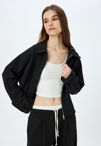 Giacca nera con zip, dal design strutturato, colletto ampio e texture a coste, abbinata a un top corto bianco a coste e pantaloni neri.