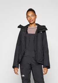 Schwarze isolierte Jacke mit abnehmbarer Kapuze, Reißverschlusstaschen und hohem Kragen. Trägt man über einem strukturierten Oberteil und passenden Latzhosen.