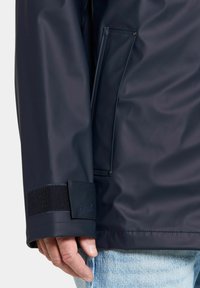 Marinblå vattentät jacka med slät yta, utrustad med kardborreärm, logotypmärke och en sidoficka på bröstet. Texturerat inre.