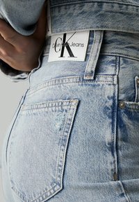 Šviesiai mėlynos džinsinės kelnės su aukštu liemeniu, su Calvin Klein Jeans etikete, matomomis siūlėmis ir nenaudotu užpakaliniu kišenės detaliu.