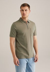 Polo de color verde oliva con cuello corto de cremallera, bolsillo en el pecho y mangas cortas, hecho de una tela suave y ligera. Ajuste casual.