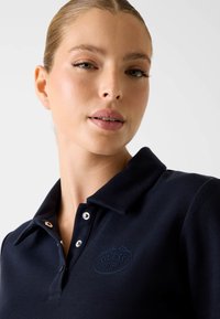 Granatowa koszulka polo z fakturowanej tkaniny, kołnierzykiem i dwoma srebrnymi guzikami. Na piersi znajduje się subtelne haftowane logo.