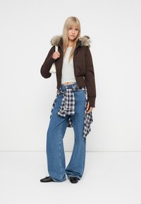 Hnědá péřová bunda s umělou kožešinou na kapuci, šedý zkrácený top, oversized modré džíny a kostkovaná košile uvázaná kolem pasu. Černé boty a bílá taška.