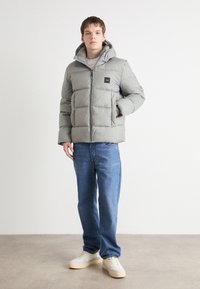 Lysgrå pufferjakke med hætte, der har lynlås foran, to sidelommer og et lille sort logo patch på brystet. Kombinationen består af blå jeans og beige sneakers.