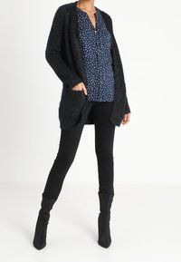 Czarny puszysty kardigan z otwartym przodem, noszony na granatowej koszuli w białe kropki, zestawiony z dopasowanymi czarnymi spodniami i wysokimi kozakami za kolano.