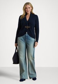 Sieviete valkā tumši zilu kabeļadījuma kardiganu ar sprādzi, gaiši zilas platu kāju džinsas, tumši zilas kurpes un tur tumši zilu rokassomiņu.