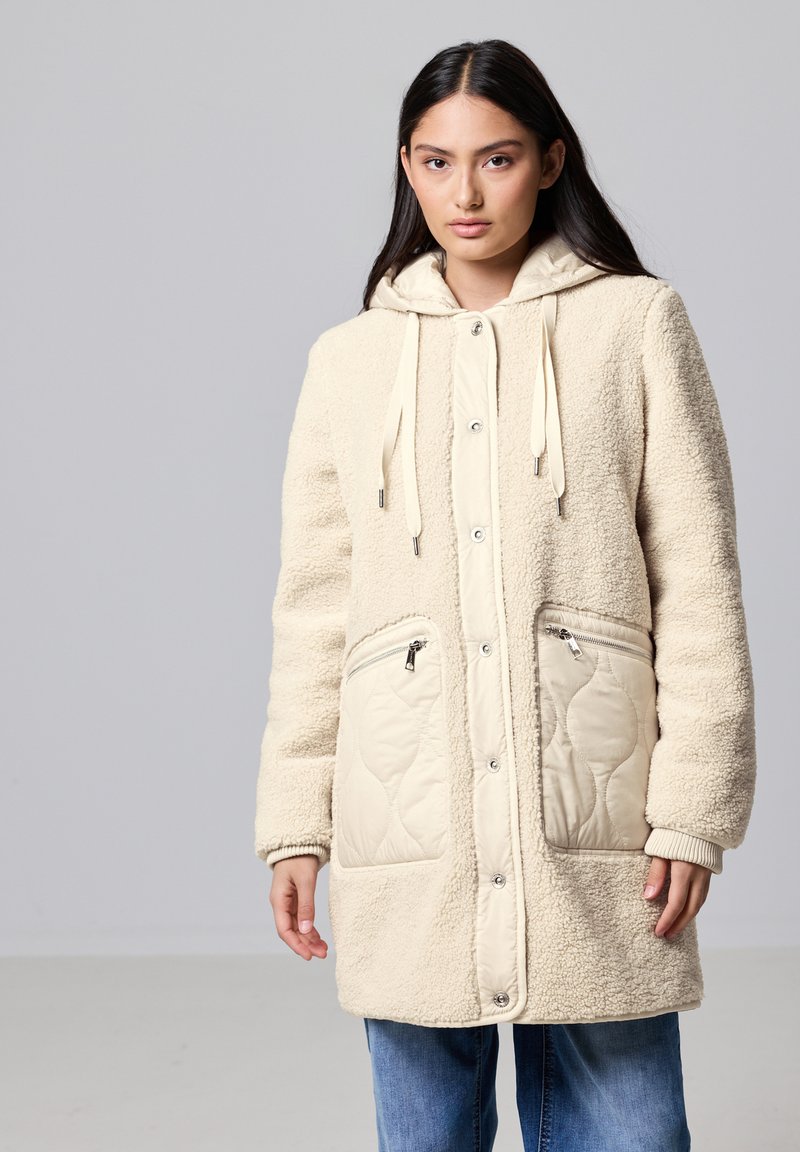 Abrigo de forro polar color crema con un frente de tela acolchada, bolsillos con cremallera y capucha ajustable. Presenta cierres de broche y puños de canalé.
