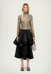 Gold sequined turtleneck top, black layered ruffled skirt, strappy black sandals, holding a small light gray handbag. Simple background.