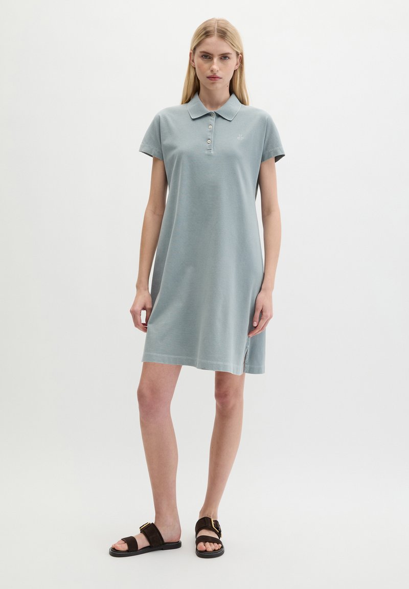 Femme aux cheveux blonds et raides porte une robe polo bleu clair à manches courtes et des sandales noires à boucle, debout sur un fond blanc uni.