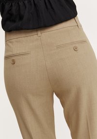 Pantaloni beige sartoriali realizzati in tessuto morbido, con due tasche posteriori con chiusura a bottone e una texture sottile.