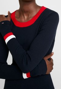 Pull côtelé bleu marine avec un large col rouge. Les manches texturées présentent des accents rouges, blancs et bleus aux poignets. Coupe ajustée.