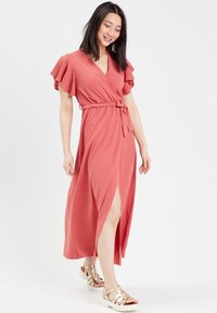 Robe portefeuille corail avec des manches volantes, une ceinture à nouer et une fente haute jusqu'à la cuisse. Assortie avec des sandales métalliques. Texture douce, design fluide.