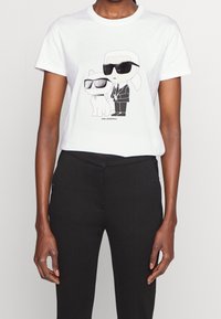 Vitt bomullströja med en svart grafisk illustration av en karaktär och en katt, båda iförda solglasögon, framtill.