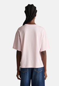 Tílko s krátkým rukávem v pastelově růžové barvě s kulatým výstřihem, volného střihu a hladkou bavlněnou texturou, v kombinaci s modrými džínami.