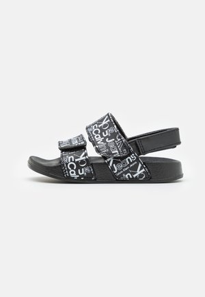 Sandalia negra con tres correas ajustables que presentan el texto del logotipo blanco de Calvin Klein Jeans sobre una suela texturizada.