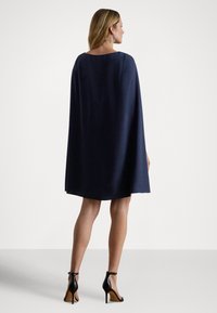 Tumši zila apmetņa kleita, ceļam līdzīga, ar gludu tekstūru, kvadrātveida izgriezumu un plūstošu dizainu. Valkāta kopā ar melnām augstpapēžu kurpēm.