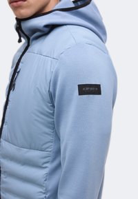 Personne portant une veste matelassée bleu clair avec une capuche et une fermeture éclair noire, montrant un patch logo noir sur la partie supérieure du bras.