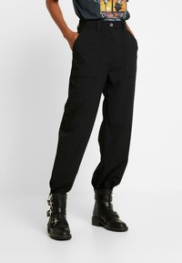 Schwarze, taillierte Hose mit hoher Taille, ausgestattet mit zwei Fronttaschen und einem Knopfverschluss, kombiniert mit schwarzen Schnallenstiefeletten.