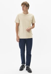 T-shirt en coton beige à manches courtes, col rond, associée à un jean bleu foncé et des chaussures noires à enfiler. Design simple, sans motifs.