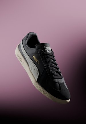 Schwarzer Sneaker mit grauen Akzenten, der ein strukturiertes Obermaterial und eine kontrastierende cremefarbene Gummisohle aufweist. Schnürsenkel und Branding auf der Zunge sichtbar.