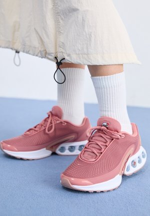 Pieds portant des chaussettes blanches et des baskets roses avec amorti à air visible, debout sur un sol bleu, ourlet de jupe au-dessus des chevilles.