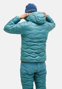 Teal isoleret pufferjakke med teksturerede bølgemønstre, hætte og elastiske manchetter. Båret med matchende bukser, der viser et sammenhængende look.