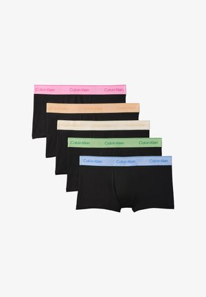 Muskulöser Mann trägt schwarze Boxershorts mit pinkem Calvin Klein-Bund und steht barfuß vor einem schlichten grauen Hintergrund.