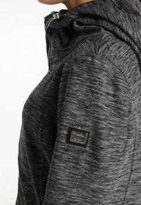 Gros plan d'une veste zippée grise mouchetée avec capuche et un patch logo noir rectangulaire sur le haut de la manche.