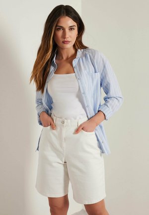 Chemise boutonnée rayée bleu clair sur un débardeur blanc, associée à un short en jean blanc. La chemise comporte une poche avant et des manches retroussées.