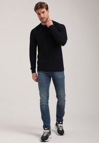 Dunkelblauer Strickpullover mit Rundhalsausschnitt, kombiniert mit hellblauen Jeans und schwarzen Sneakern. Der Pullover hat ein strukturiertes Muster und lange Ärmel.