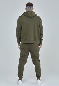 Conjunto de sudadera y pantalones jogger en verde oliva, hecho de una tela suave, con un diseño sencillo y un corte clásico, y puños acanalados. Incluye zapatillas blancas.