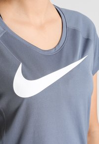 Camiseta deportiva de manga corta gris hecha de tela suave, con un gran logo blanco de Nike en forma de swoosh en el frente. Cuello redondeado.