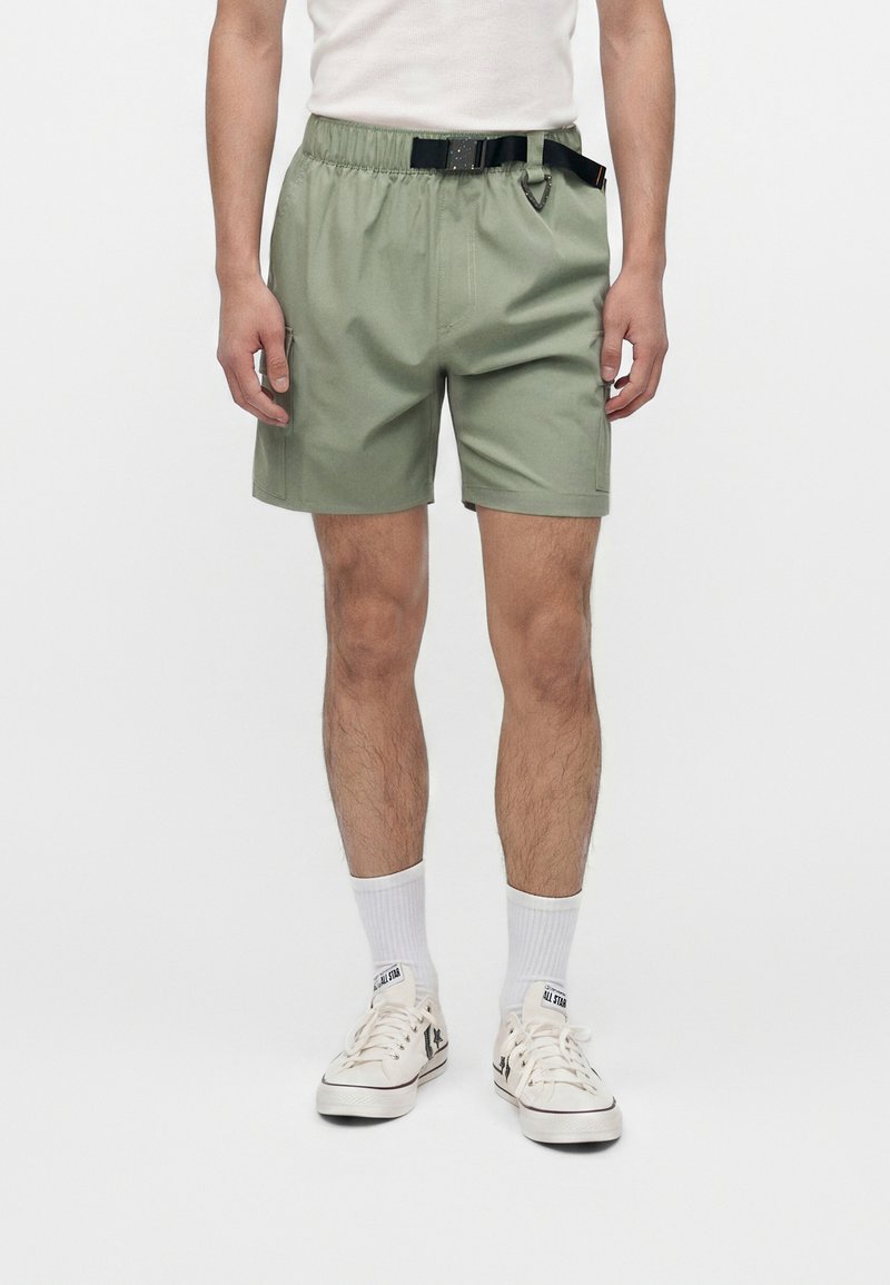 Άνδρας που φορά ανοιχτοπράσινα σορτς με ενσωματωμένη μαύρη ζώνη, λευκές κάλτσες και λευκά χαμηλά αθλητικά παπούτσια, στέκεται σε απλό φόντο.