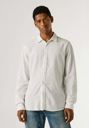 Mladý muž s krátkymi kučeravými vlasmi, oblečený v bielej košeli s dlhým rukávom a modrých rifliach, stojaci pred jednoduchým pozadím.