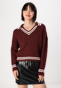 Maglione in cotone borgogna a lavorazione a trecce con scollatura a V e colletto a righe bianche; abbinato a una mini gonna nera in pelle di coccodrillo sintetica con cintura.