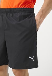 Melni sporta šorti, kas izgatavoti no viegla auduma, ar elastīgu jostu, sānu kabatām un baltu Puma logo kreisajā apakšējā kājā.